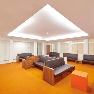 Großzügiger Aufenthaltsbereich in einem Bürogebäude in der Marburger Straße in Berlin mit Sitzmöbeln aus Holz und Polster, orangefarbenem Teppichboden und indirekter Deckenbeleuchtung.