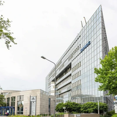 Außenansicht des Delta Haus in Wiesbaden mit moderner Glasfassade und umliegender Begrünung an einer Straße.