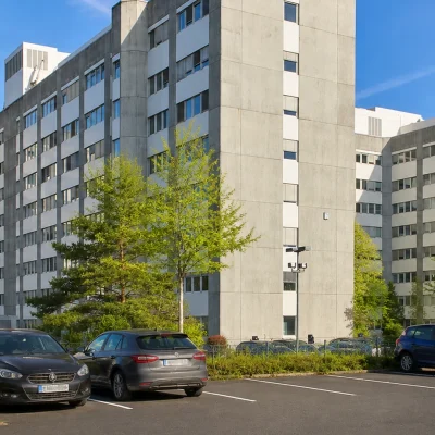 Bürogebäude mit markantem Treppenhausturm, Parkplatz und Bäumen im Frühlingslaub