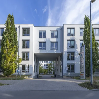 Weißes Bürogebäude mit Tordurchfahrt, Säulenpappeln und Firmenschild an der Zufahrt