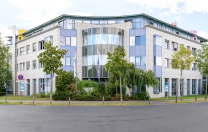 Bürogebäude mit runder Glasfront, Natursteinfassade und Grünanlage bei bewölktem Himmel