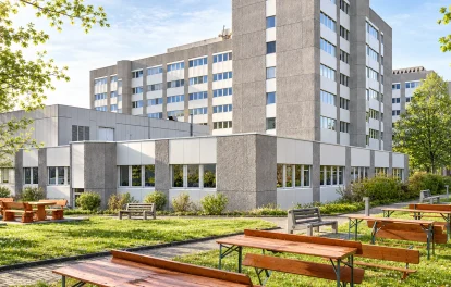 Bürokomplex mit Betonfassade hinter Frühlingsbäumen, davor Wiese mit Biertischgarnituren