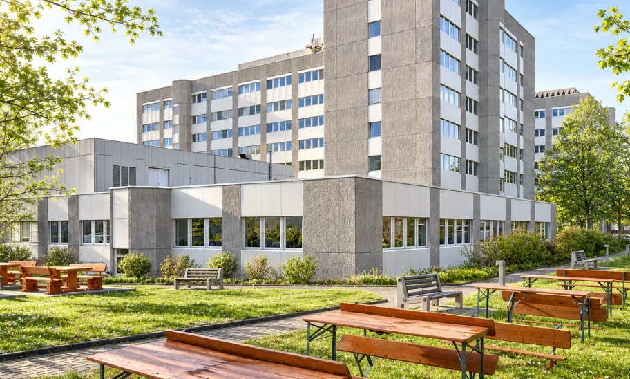Bürokomplex mit Betonfassade hinter Frühlingsbäumen, davor Wiese mit Biertischgarnituren