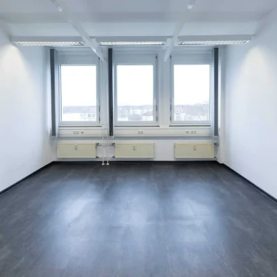 Leerer Büroraum mit drei Fenstern, hellen Wänden und dunklem Vinylboden
