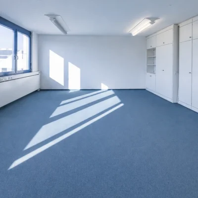 Leeres Großraumbüro mit blauem Teppichboden, Fensterfront und weißen Einbauschränken
