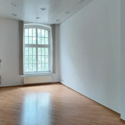 Leerer Büroraum mit Rundbogenfenster, hellem Holzboden und Deckenstrahlern