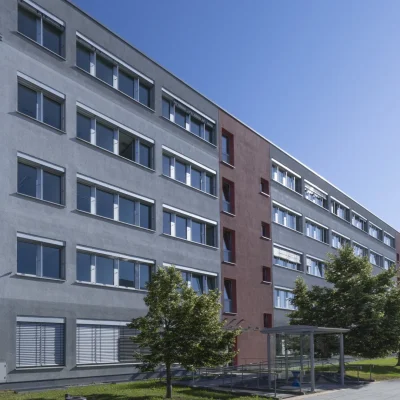 Gebäudeecke mit grauer und roter Fassade sowie Bäumen davor