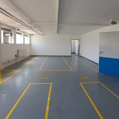 Leere Lagerhalle mit Fenstern, gelber Bodenmarkierung und blauer Tür