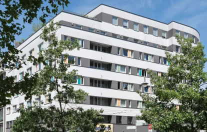 Weißes Hotelgebäude mit grauen Fensterbändern und Bäumen im Vordergrund an einer sonnigen Berliner Straßenecke