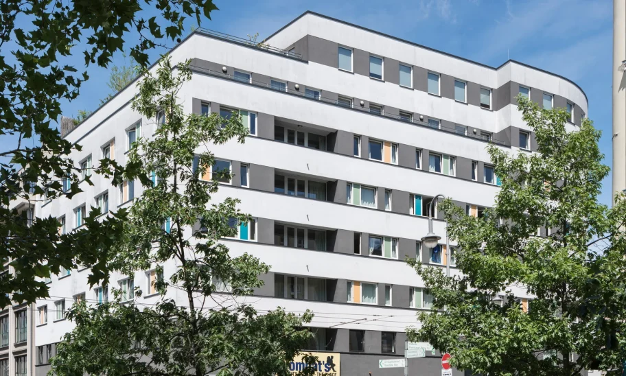 Weißes Hotelgebäude mit grauen Fensterbändern und Bäumen im Vordergrund an einer sonnigen Berliner Straßenecke