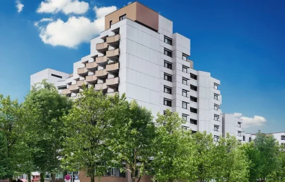 Terrassenartig gestaffeltes Bürogebäude mit heller Fassade und Balkonen hinter Straßenbäumen und Parkplatz