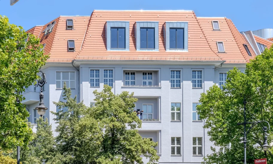 Berliner Altbau mit rotem Ziegeldach, Zinkgauben und Straßenbäumen vor blauem Himmel