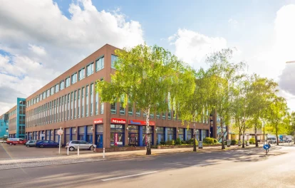 Bürogebäude mit Ladenzone im Erdgeschoss an einer Straßenecke, davor Birken unter blauem Himmel
