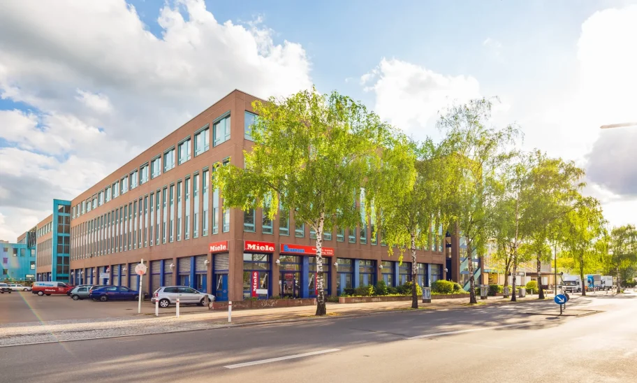 Bürogebäude mit Ladenzone im Erdgeschoss an einer Straßenecke, davor Birken unter blauem Himmel