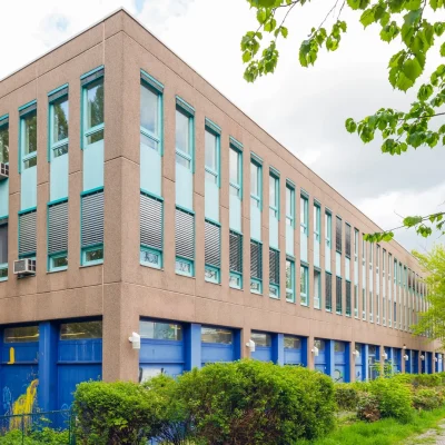 Zweigeschossiges Bürogebäude mit türkisen Fensterrahmen und blauem Sockel, umgeben von Grün an der Straße