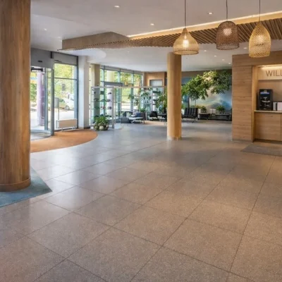 Großzügiges Foyer mit Holzsäulen, Sitzecken, Pendelleuchten und Empfangstresen mit Willkommen-Schriftzug (KI-Visualisierung)