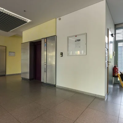 Eingangsbereich mit Aufzug, Wegweiser Erdgeschoss, Zimmerpflanze und Steinboden vor großen Fenstern