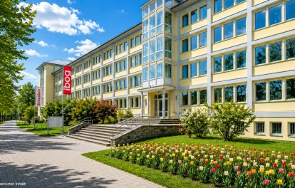 Gelbes Bürogebäude mit Eingangstreppe, Tulpenbeet, Fahnen und Gehweg unter blauem Himmel (KI-Visualisierung)