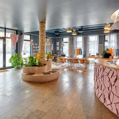 Lobby mit geschwungener rosa Rezeption, lichterumwickelter Säule mit Pflanzen und großen Fenstern zur Straße