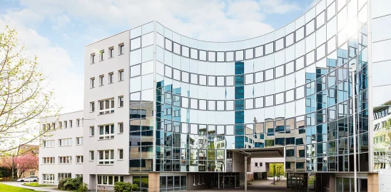 Frontale Außenansicht eines modernen Büroensembles in der Heßbrühlstraße in Stuttgart mit markanter, gebogener Glasfassade, die den Vorplatz sowie umliegende Gebäude widerspiegelt; helle, klare Architektur unter leicht bewölktem Himmel.