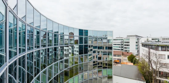 Geschwungene Glasfassade eines modernen Bürogebäudes in der Heßbrühlstraße in Stuttgart, die umliegende Gebäude und den Himmel spiegelnd reflektiert; Blick auf den Innenhof und angrenzende Architektur.