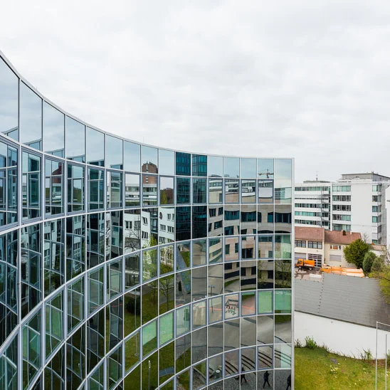 Geschwungene Glasfassade eines modernen Bürogebäudes in der Heßbrühlstraße in Stuttgart, die umliegende Gebäude und den Himmel spiegelnd reflektiert; Blick auf den Innenhof und angrenzende Architektur.