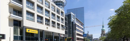 Das Bild zeigt das Westfalenforum Dortmund aus der Straßenperspektive mit U-Bahn-Zugang, Einkaufsmöglichkeiten und Grünfläche im Vordergrund.